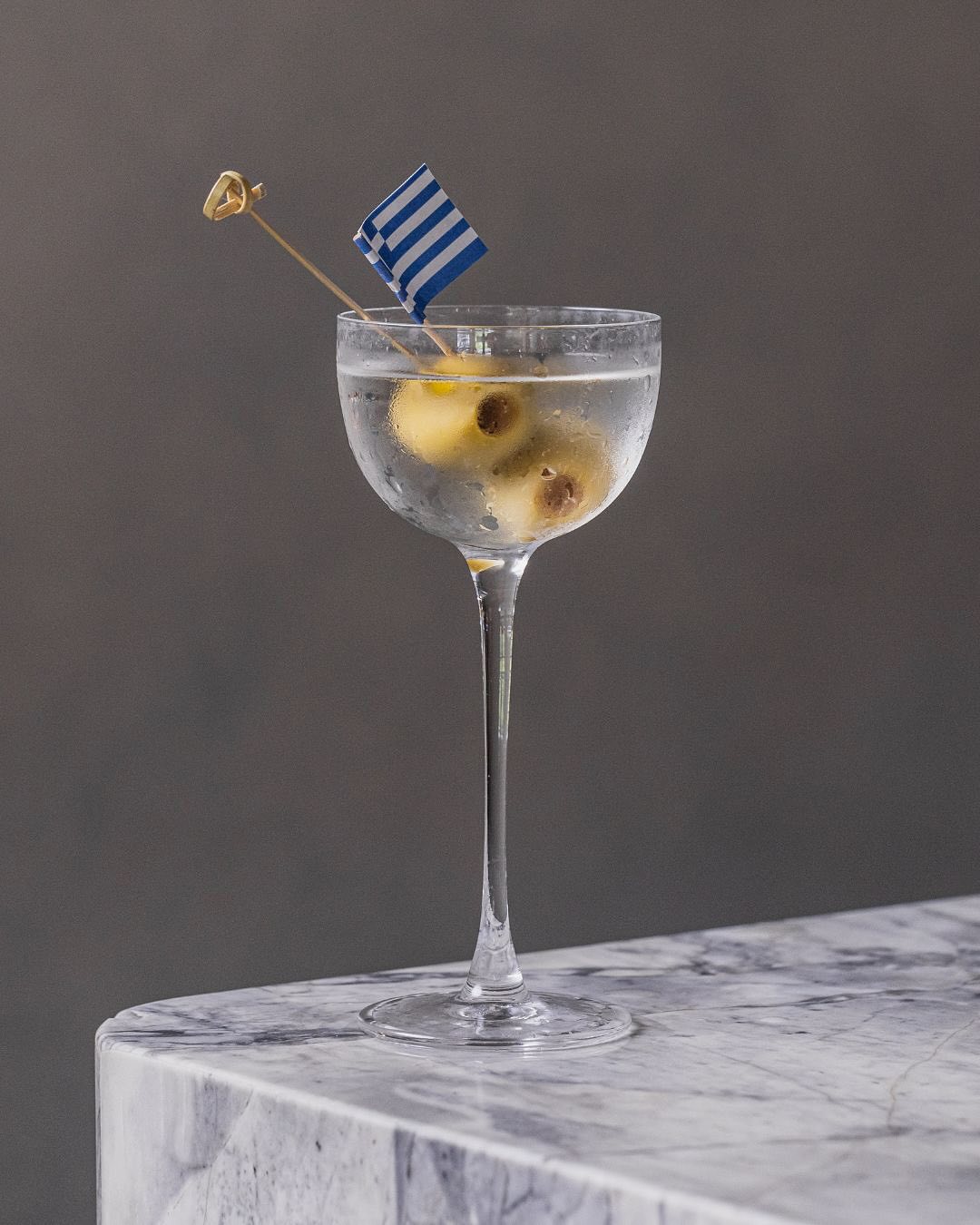A gin Martini garnished with olives and a Greek flag.